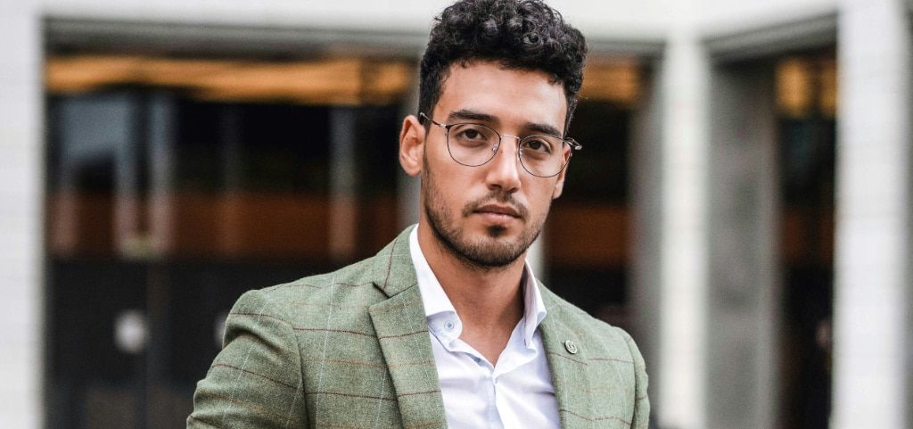 A young man with closely-cropped curly black hair and light stubble. He is wearing a tweed jacket and round metal glasses. He appears to be standing in a modern cityscape which is blurred in the background.