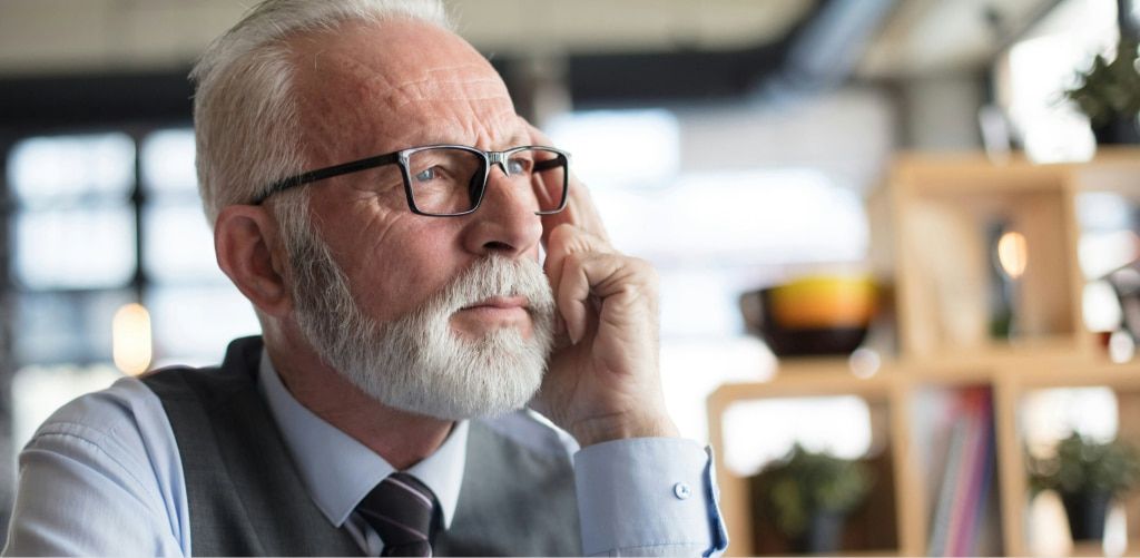 An older man with short white hair and a full white beard wears black rectangular glasses while sitting indoors in a modern café setting. Dressed in a light blue shirt, tie, and grey waistcoat, he rests his face against his hand and gazes thoughtfully int