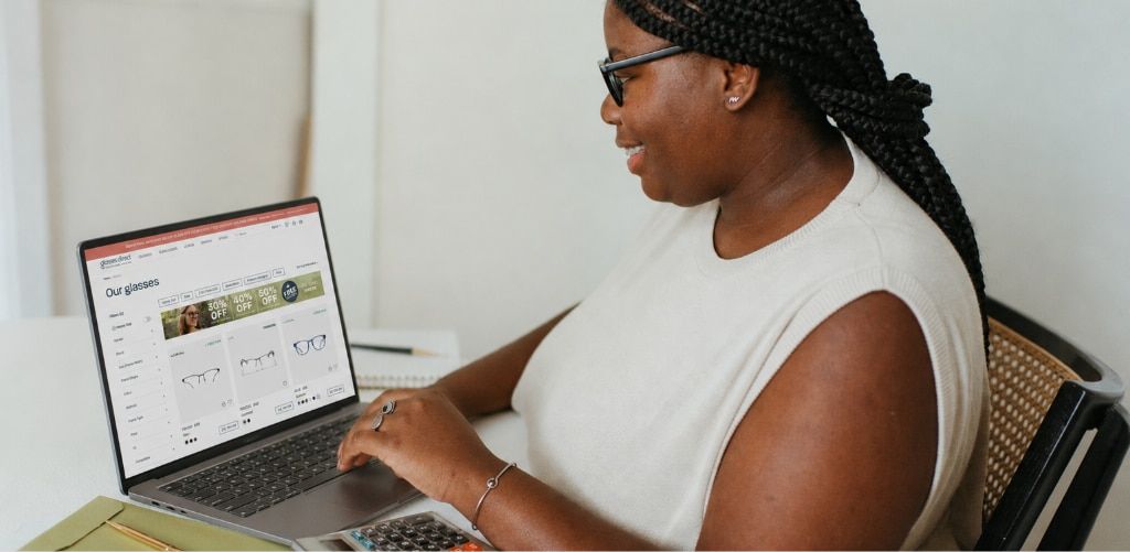 A woman with braided hair, wearing dark acetate glasses and a white sleeveless top, sits at a desk using a laptop to browse Glasses Direct's online eyewear store. The screen shows a selection of eyeglass frames and filtering options, while a calculator an