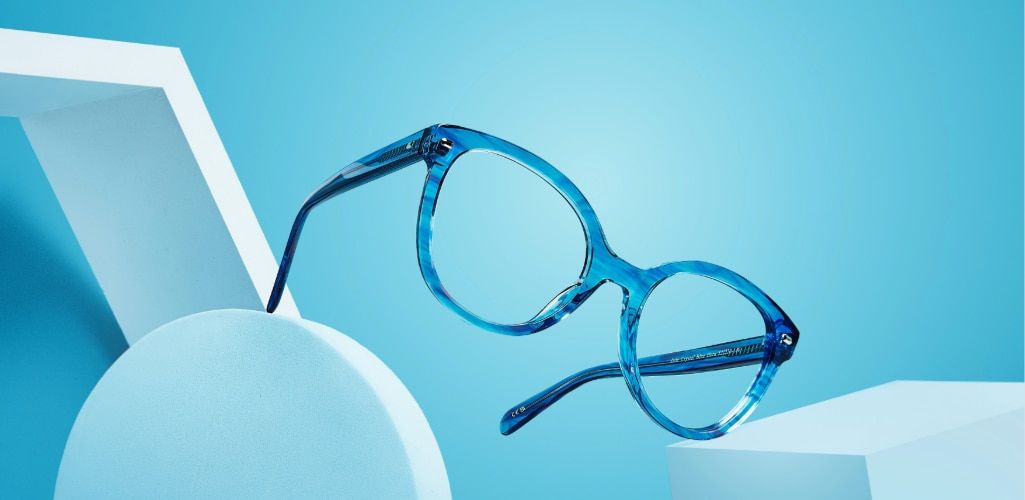 Creative product photograph of Scout Jade oversized translucent blue glasses suspended above geometric display props against a bright blue background. The frame features large rounded cat-eye lenses, a thick acetate construction and a glossy finish that c