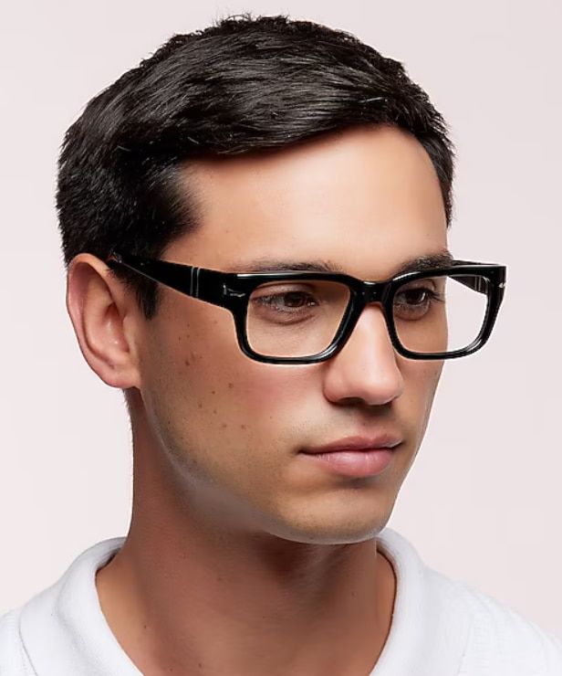 A man with neatly combed dark hair wears a white polo shirt and bold black rectangular glasses while looking downward and slightly to the side. Set against a soft neutral background, the portrait highlights the thick acetate frames and gives the image a p