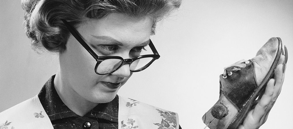 A black-and-white close-up image of a lady with 50s-style clothing and a short perm. She is wearing a pair of dark thick-rimmed glasses which sit on the edge of her nose. She appears to be inspecting and cleaning a small shoe which she is holding up to he