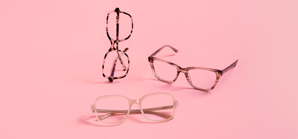 Three Pink Ribbon acetate glasses are displayed against a pastel pink background. One pair, a pink Havana square frame, stands upright on its side. Beside it rests a pink and brown striped cat-eye frame, while in front, a nude square frame sits neatly fol