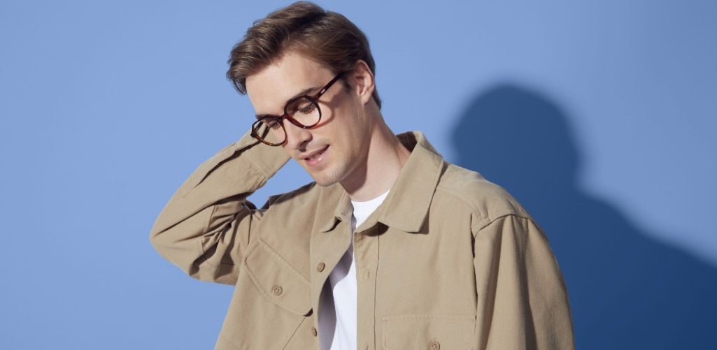 Model wearing round tortoiseshell Levi's eyeglasses and a beige jacket, posing against a blue background with his hand behind his head.