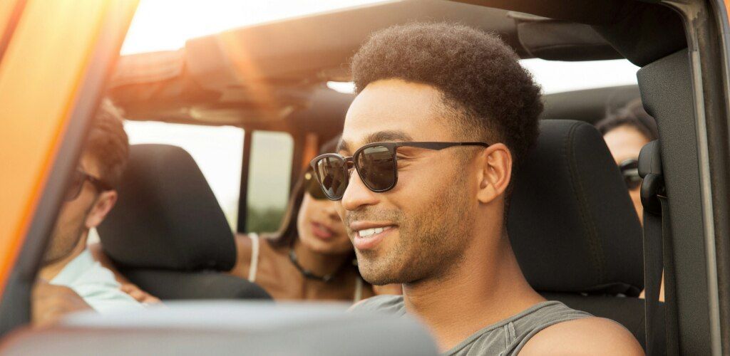 Man with short afro wearing black square prescription sunglasses driving in an open-top car in bright sunshine with friends.