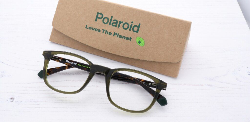 A pair of olive-green Polaroid sustainable eyeglass frames is displayed on a white wooden surface beside a kraft-paper glasses case printed with the message “Polaroid Loves The Planet.” The glasses feature a rounded-square shape with translucent green rim