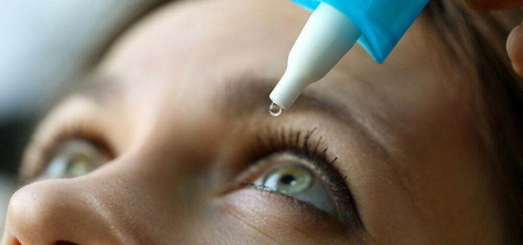 A close-up of a woman applying eye drops to her left eye. She has light brown hair and striking green eyes.