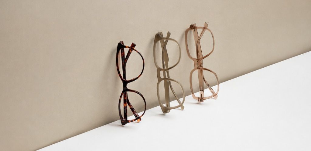 Three pairs of Glasses Direct frames are displayed upright against a soft beige backdrop. The frames feature different finishes—a Havana pattern, a muted matte green and a translucent shiny beige—arranged on a clean white surface. The minimalist compositi