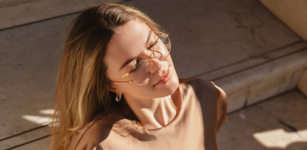 Woman with long blonde hair wearing round gold metal glasses in warm sunlight, with a minimalist neutral outfit.