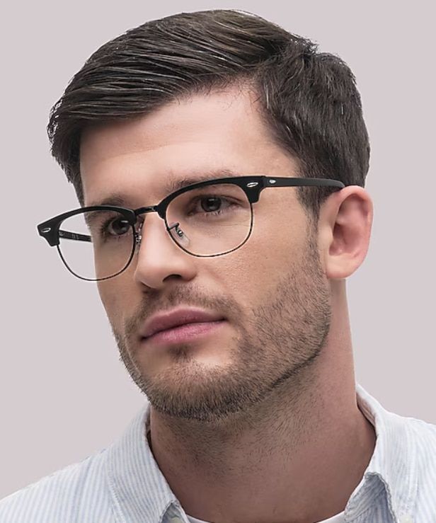 A man with neatly styled dark hair and light stubble wears a white pinstripe shirt and black Clubmaster-style glasses while looking slightly off-camera. Set against a clean neutral background, the portrait highlights the glasses’ bold upper frame and thin