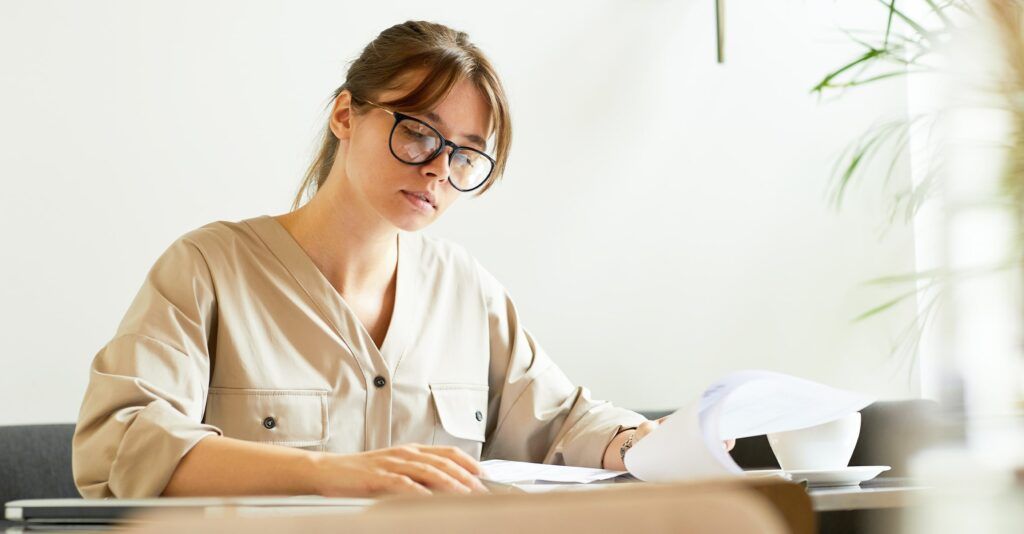 Woman with brown hair tied in a ponytail wearing black angular glasses, and a beige shirt, seated at a desk reviewing paperwork in a bright, modern kitchen setting.