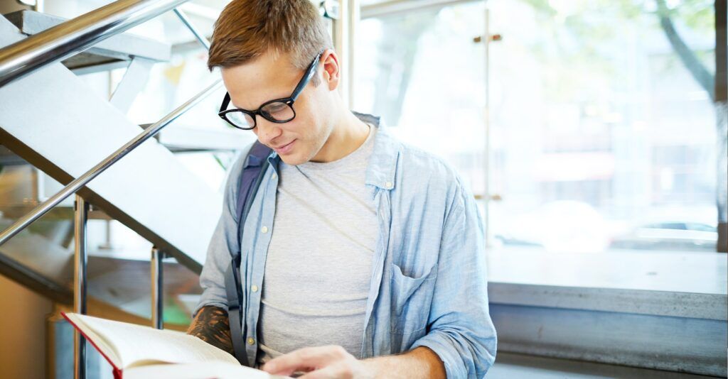 Man with short blonde hair, wearing black Wayfarer frames and a blue shirt over a white tee, reading a book in a bright library setting.