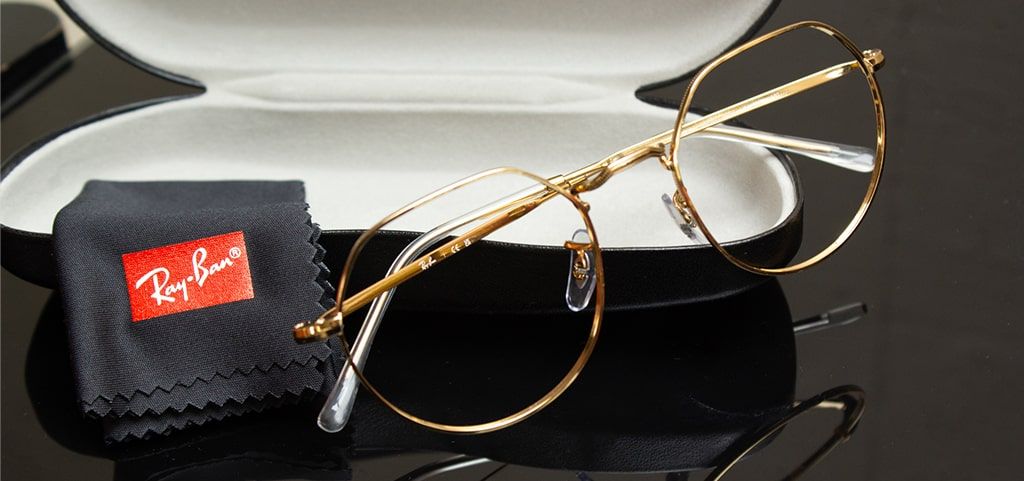 A close-up shot of a folded gold geometric metal frame resting on a black Ray-Ban glasses case, accompanied by a neatly folded Ray-Ban cleaning cloth beside it. The background is a sleek, glossy black surface.