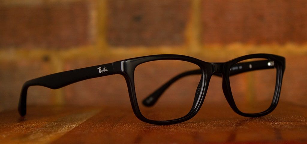 An open pair of black acetate Ray-Ban glasses positioned at an angle on a wooden surface, with a blurred backdrop of a brick wall.