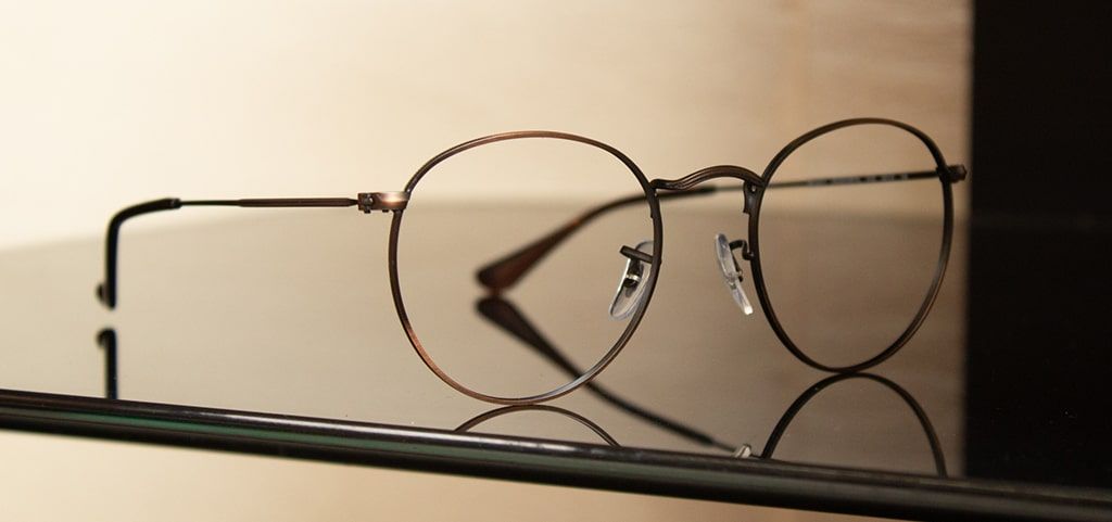A pair of round gold metal glasses positioned at an angle on a glossy black surface, the backdrop is lit slightly, casting shadows.