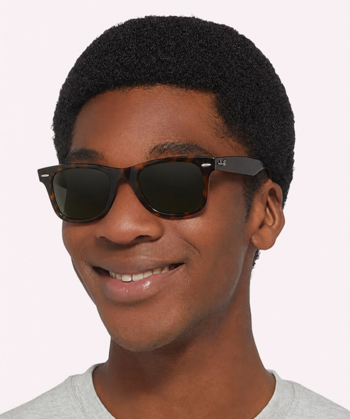A smiling man with a short black afro wears dark Havana Ray-Ban Wayfarer sunglasses and a grey t-shirt, looking slightly to the side against a plain background.