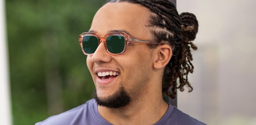 Close-up of Perri Kiely smiling brightly with his hair tied back, wearing a blue t-shirt and Ray-Ban sunglasses with translucent pink frames and green-tinted lenses, standing outdoors in bright sunlight.