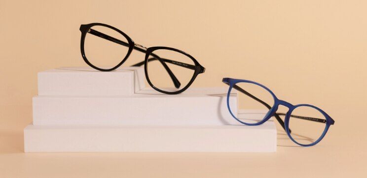Two pairs of GD Collection round acetate glasses displayed on white geometric blocks — one in shiny black, one in bright blue — against a warm neutral background