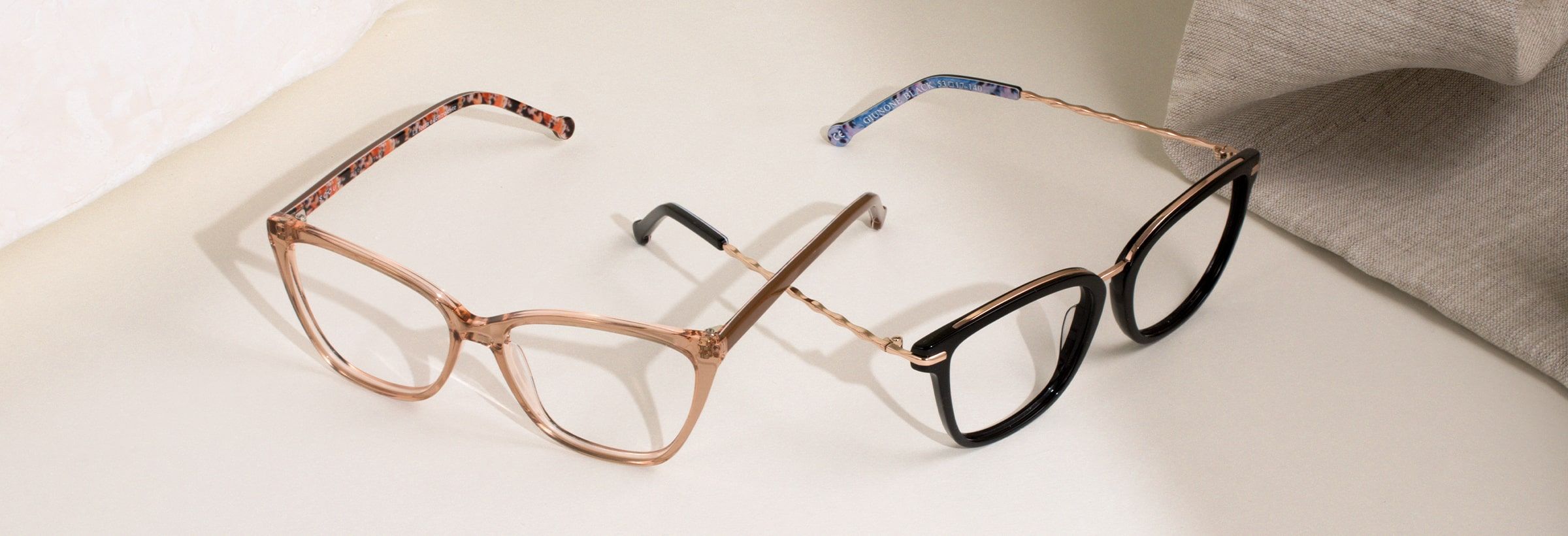 Two pairs of Scout Made in Italy frames, one beige and one black positioned facing opposite each other with arms crossed with stone blocks and fabric in the background