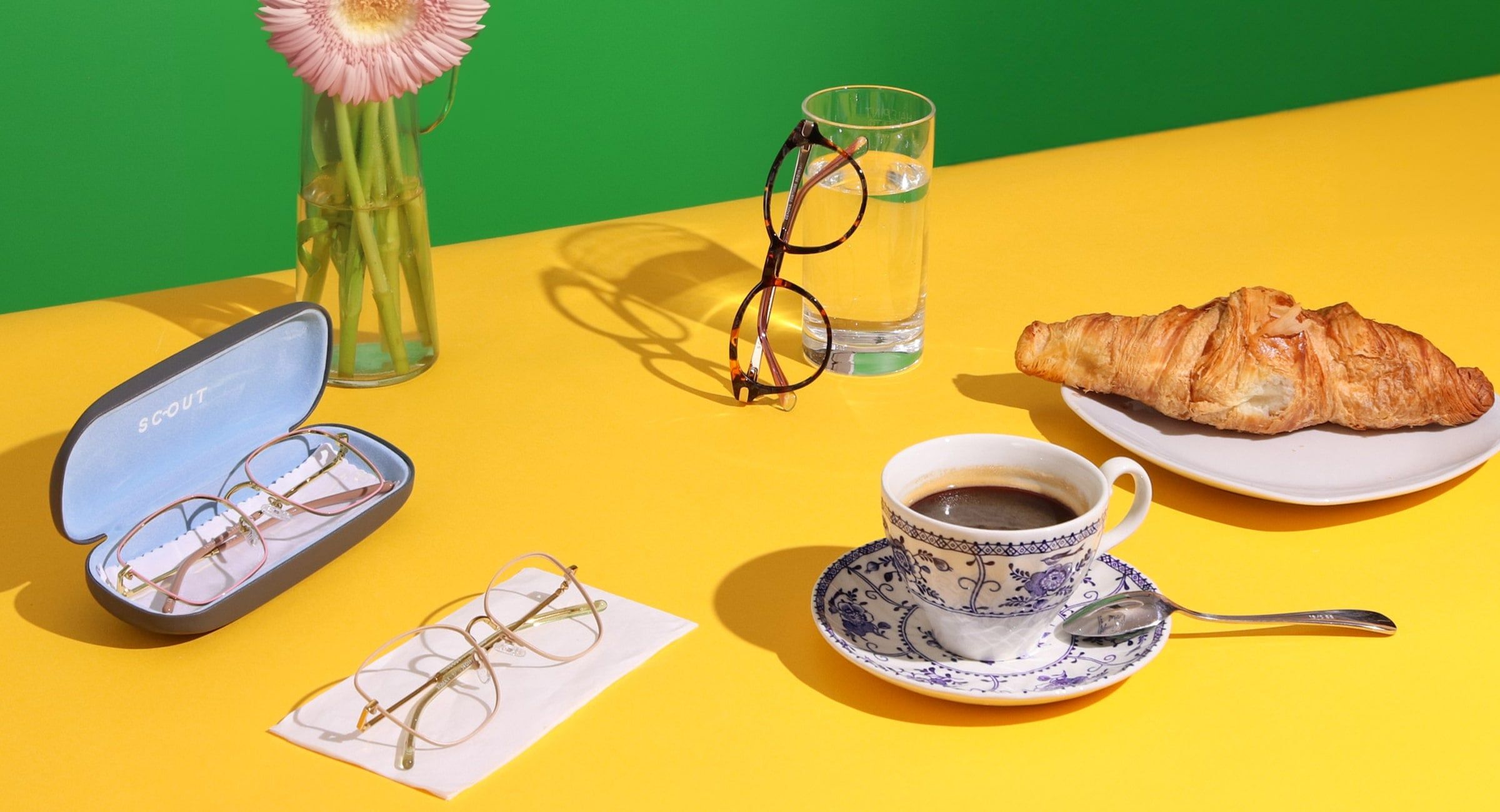 3 pairs of Scout: Made in Italy frames on a yellow and green background, one frame in its hardcase, one on a napkin and one balancing against a glass surrounded by a cup of coffee on a saucer and croissant on a plate