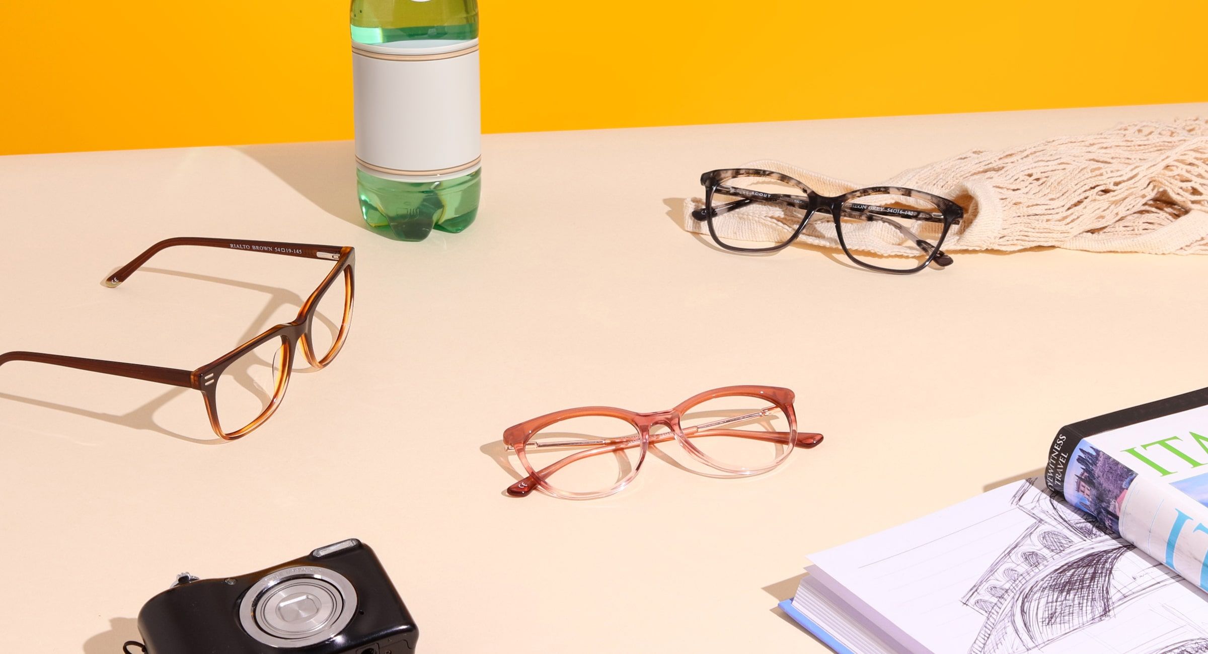 3 pairs of Scout: Made in Italy frames on a beige and yellow background surrounded by a bottle of water, string bag, digital camera, notepad and italian guide book