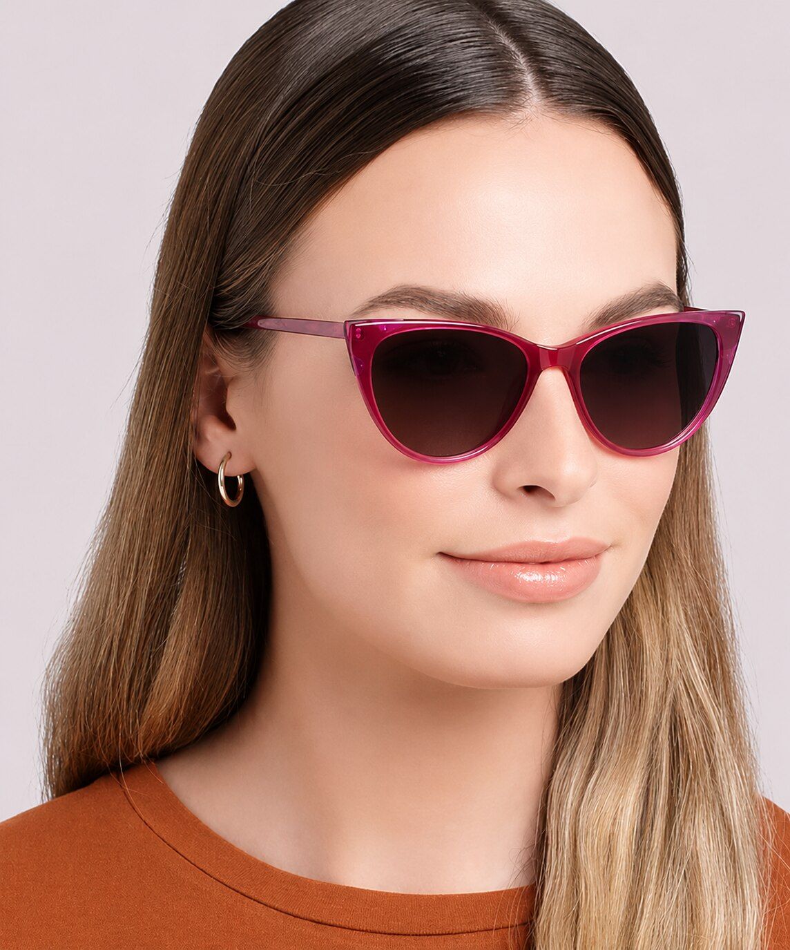 A woman with long light-brown hair wears pink cat-eye sunglasses, small hoop earrings, and an orange t-shirt, looking slightly to the side with a soft, neutral expression against a plain background.