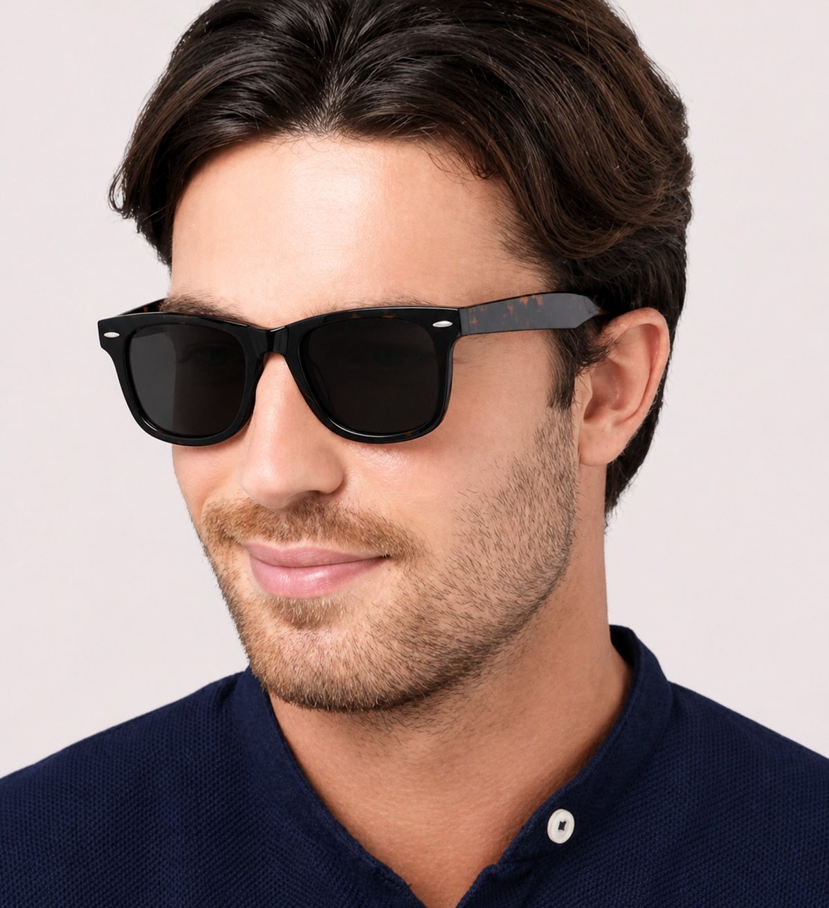 A man with dark brown, medium-length hair and light facial stubble wears Scout Costello tortoiseshell wayfarer-style sunglasses and a navy blue collared shirt with a white button. He's shown from the shoulders up against a light grey background, facing sl