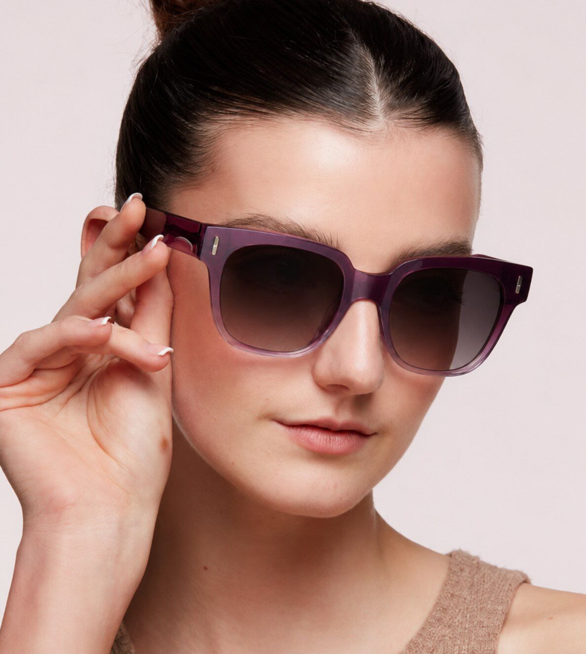 A woman with dark hair tied neatly back in a bun wears bold purple sunglasses with a light-brown vest top, gently touching the frame while looking off to the side against a plain background.