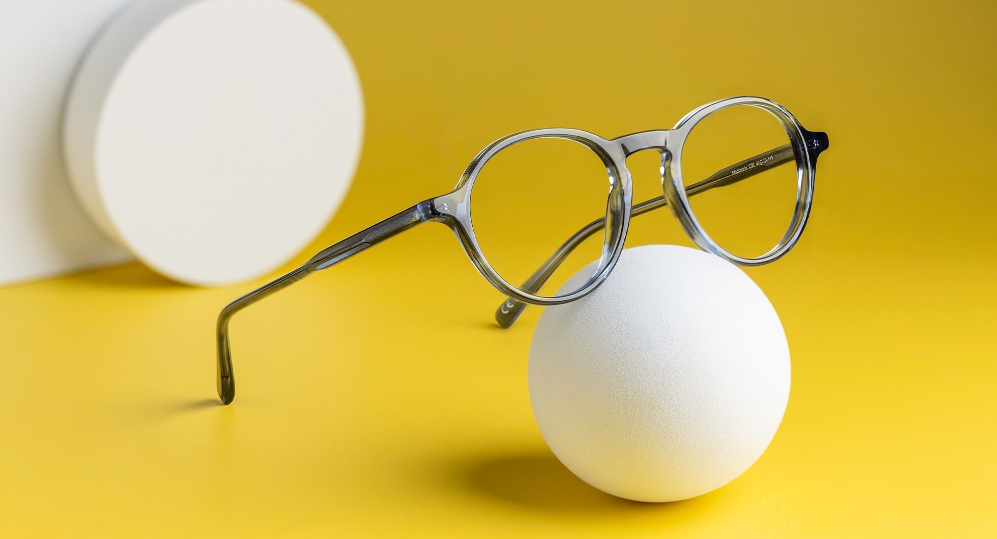 Phantos-shaped bio-acetate frames from the Scout range with a keyhole bridge, propped on a white ball, set against a bright yellow background. A white cylinder and square is just visible.