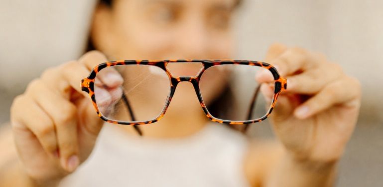 Close-up of a person holding a pair of tortoiseshell aviator glasses with visible scratches on one lens. The scratched lens appears marked and hazy compared with the clearer lens, illustrating how surface scratches can affect lens clarity and visual perfo