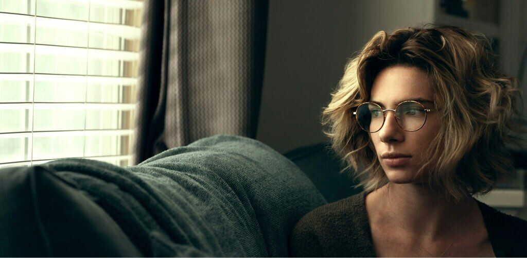 A woman with medium wavy hair wearing round metal glasses is sitting on a sofa, gazing thoughtfully at sunlight emerging through the blinds.
