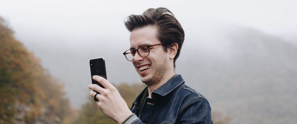 Man with a brown quiff wearing a denim jacket and round tortoiseshell acetate glasses, laughing while taking a selfie on his smartphone outdoors under grey skies