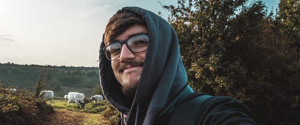 Man in grey hoodie and rectangular glasses taking a selfie outdoors, with cows and green countryside in the background.