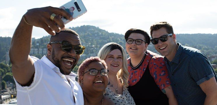 Group of five friends taking a selfie in a bright mountain setting, with a man in sunglasses in the foreground and four friends behind him, including two women wearing glasses.