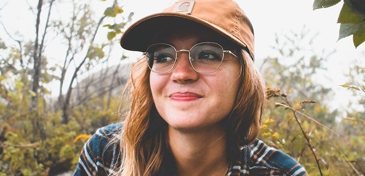 Woman wearing a brown baseball cap and check shirt with round metal glasses, looking into the distance while taking a selfie in a forest setting.