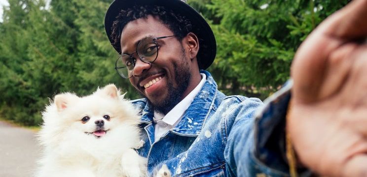 Man with short dark hair wearing a wide-brimmed hat, denim jacket, and round metal glasses, smiling while taking a selfie with his Pomeranian dog.