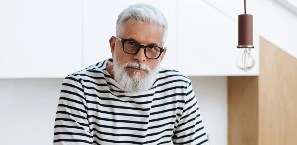 A stylish older man with white hair and a full white beard wears thick black glasses and a striped long-sleeve shirt while standing indoors in a bright modern space. He looks confidently toward the camera, with minimalist décor and a hanging light bulb ad
