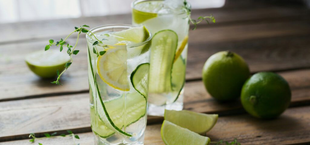 An image of two clear glass cups on a wooden surface, containing clear liquid, a slice of cucumber and a chunk of lemon and lime each. Two whole limes are visible to one side, there are also two slices and a half a lime decorating the surface.