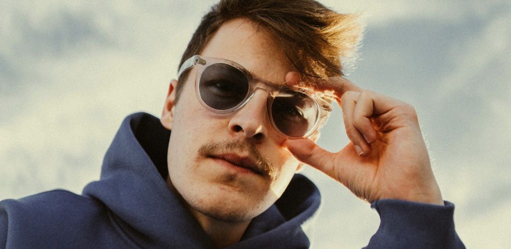 Man with light brown hair wearing sporty transparent sunglasses with a casual blue hoodie, adjusting frames outdoors in bright sunlight, styled for everyday athleisure wear.