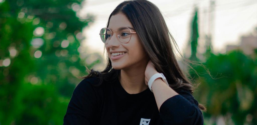 Woman with long brown hair wearing round metal glasses with a sporty black sweater, smiling outdoors in a relaxed setting, styled for everyday wear.