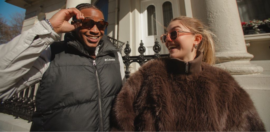 A young man in a black sleeveless puffer vest over a beige long-sleeve shirt and a young woman in a brown fluffy coat walk side by side outdoors, smiling at each other and wearing sunglasses in bright sunlight, with white columns and a black iron fence be
