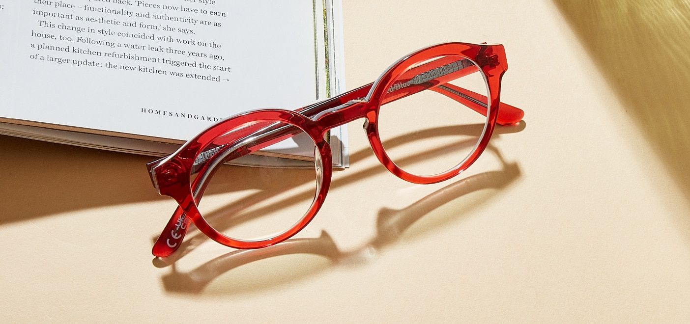 A pair of bold red, circular acetate glasses folded up and resting upwards on the edge of an open book.