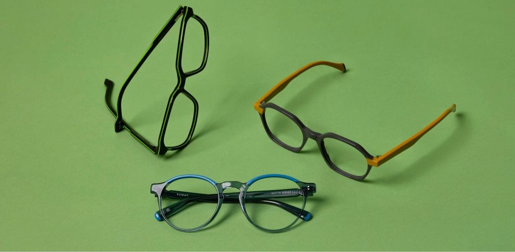 A product shot of three pairs of eyeglasses arranged on a green background. Each frame has a bold, distinctive design: one pair is black with a rectangular shape standing upright, another features a mix of grey and bright yellow accents, and the third is 