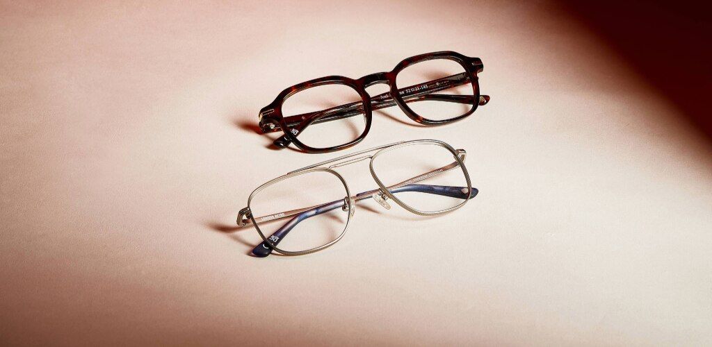 A flat lay image of two pairs of eyeglasses placed on a soft, neutral-toned surface. The top pair features bold, dark tortoiseshell square frames with a thicker, statement design, while the bottom pair has thin, light metal aviator frames in a more minima