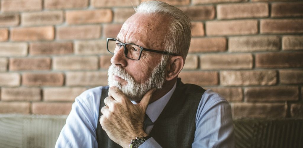 An older man with white hair and a neatly groomed white beard wears rectangular black glasses while sitting indoors against a rustic brick wall. Dressed in a light blue shirt and dark waistcoat, he rests his hand thoughtfully on his chin and looks off int