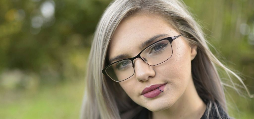 A close up of a young lady with ashy blonde long hair and grey-coloured eyes. She is wearing a pair of rectangular, black metal glasses and a black jacket. The lady has a lip piercing and is tilting her head to one side. The background is blurred but appe