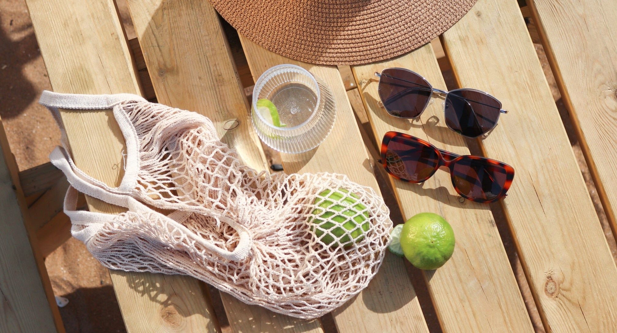 On a sunlit wooden slatted bench at the beach rests a collection of summer essentials: the brim of a hat, a netted bag, two limes and a glass of water. Beside them lie two pairs of sunglasses — one a silver geometric metal design, the other an oval Havana