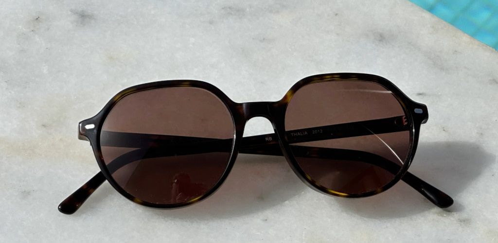 Round tortoiseshell prescription Ray-Ban sunglasses with brown lenses resting on a marble surface beside a swimming pool.