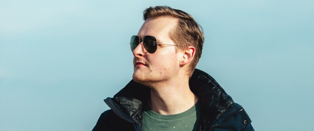 A man seen from the shoulders upwards wearing pilot-shaped sunglasses looking to the left of the screen (his right). He is wearing a black coat and green sweater. There is a sky-blue background.