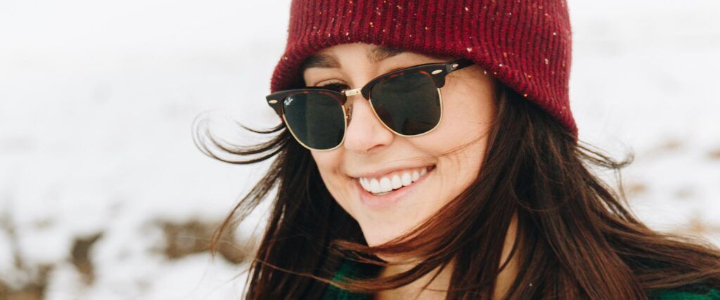 A woman with brown hair wearing a woollen red hat and Clubmaster sunglasses. She is smiling brightly. The background is snowy.